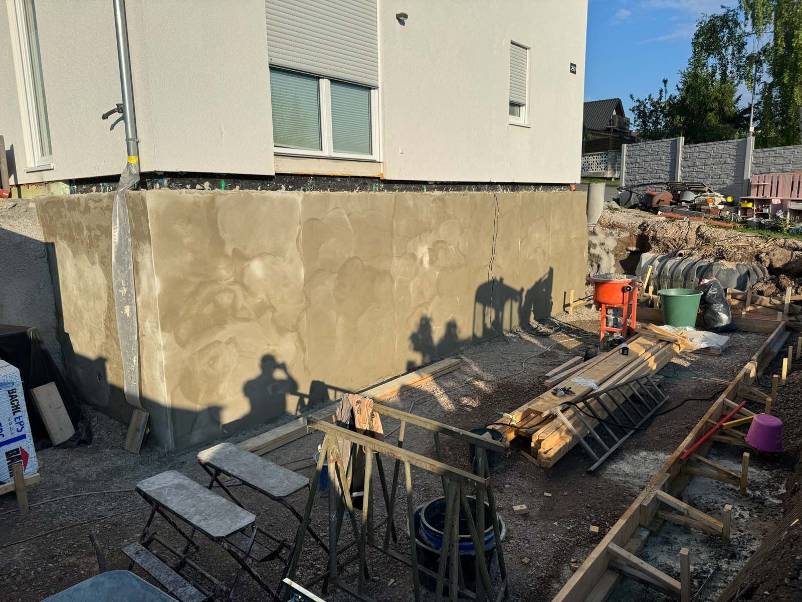 Baustelle neben einem Haus, mit nasser Betonwand, Gerüstbohlen, Werkzeugen und Baumaterialien auf schlammigem Boden