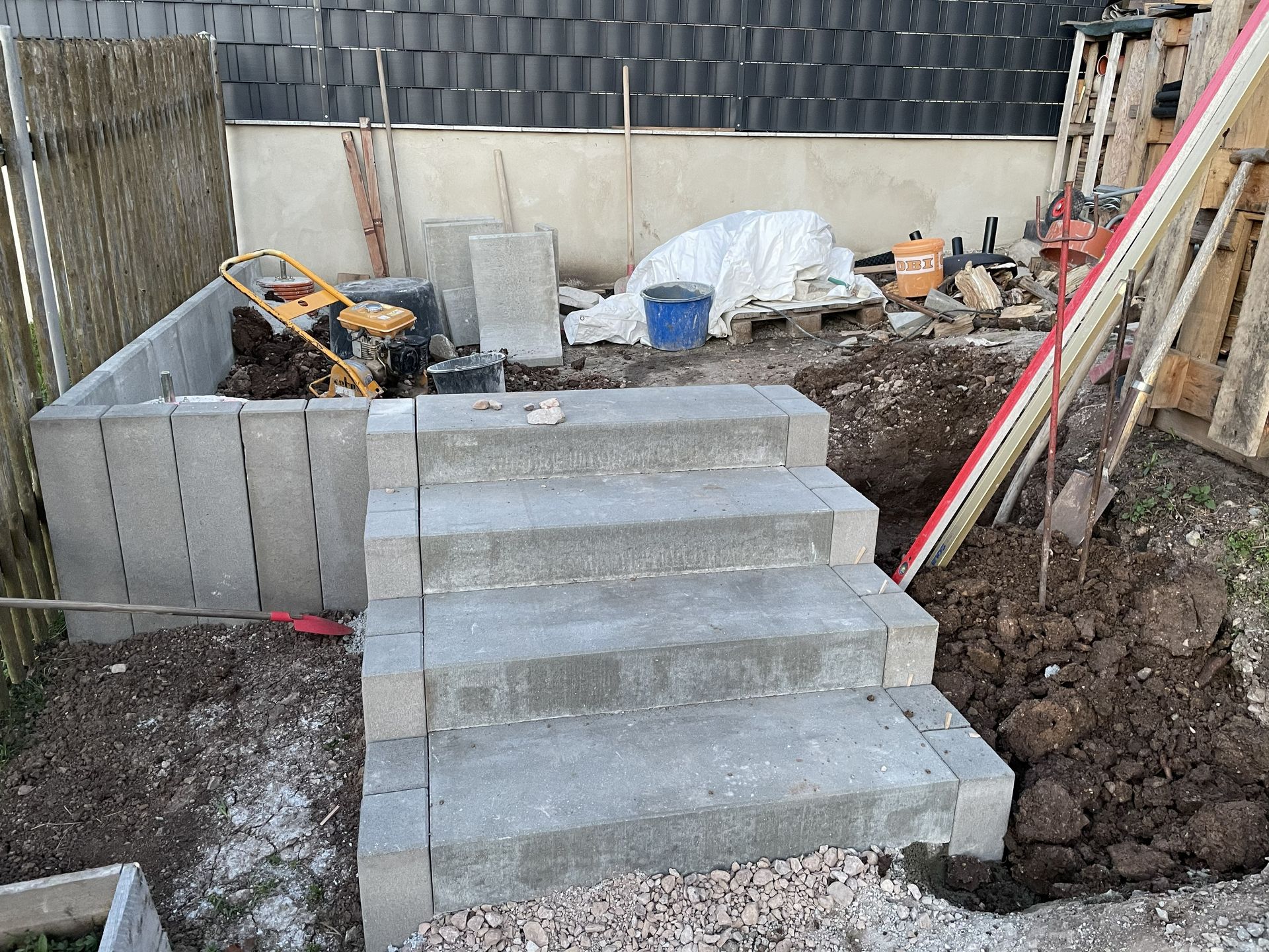 Im Bau befindliche Betontreppen in einem Hinterhof, umgeben von Erdhaufen und Werkzeugen.
