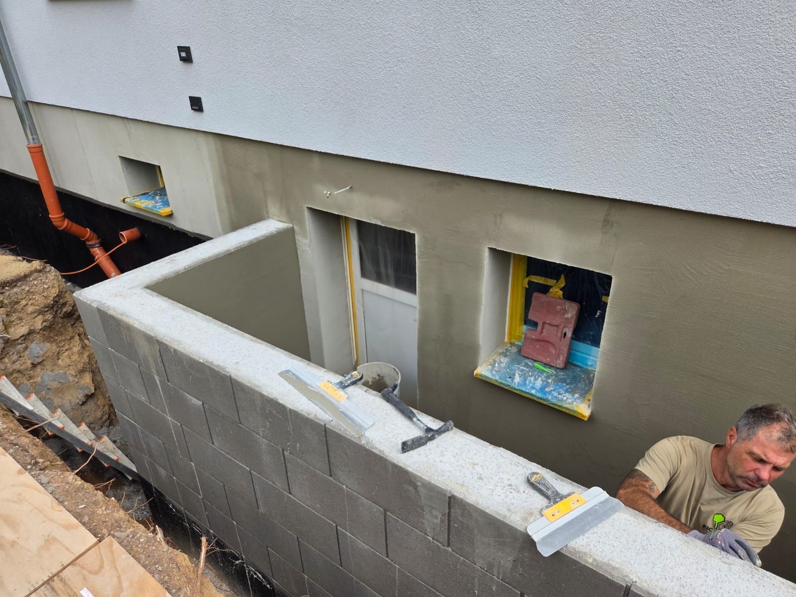 Arbeiter errichten neben einem Haus eine graue Blocksteinwand im Keller, mit offenen Lichtschächten und Baumaterialien.