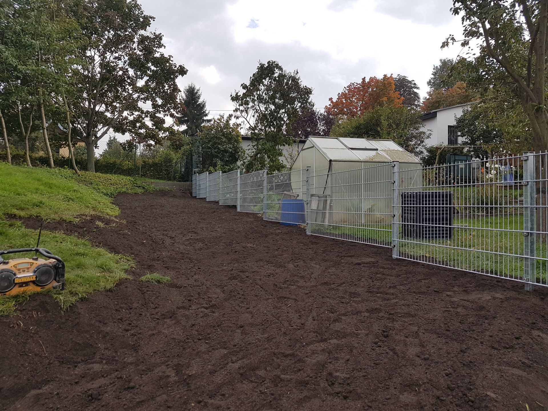 Frisch gepflügter Gartenweg neben einem Metallzaun, im Hintergrund Bäume und Häuser.