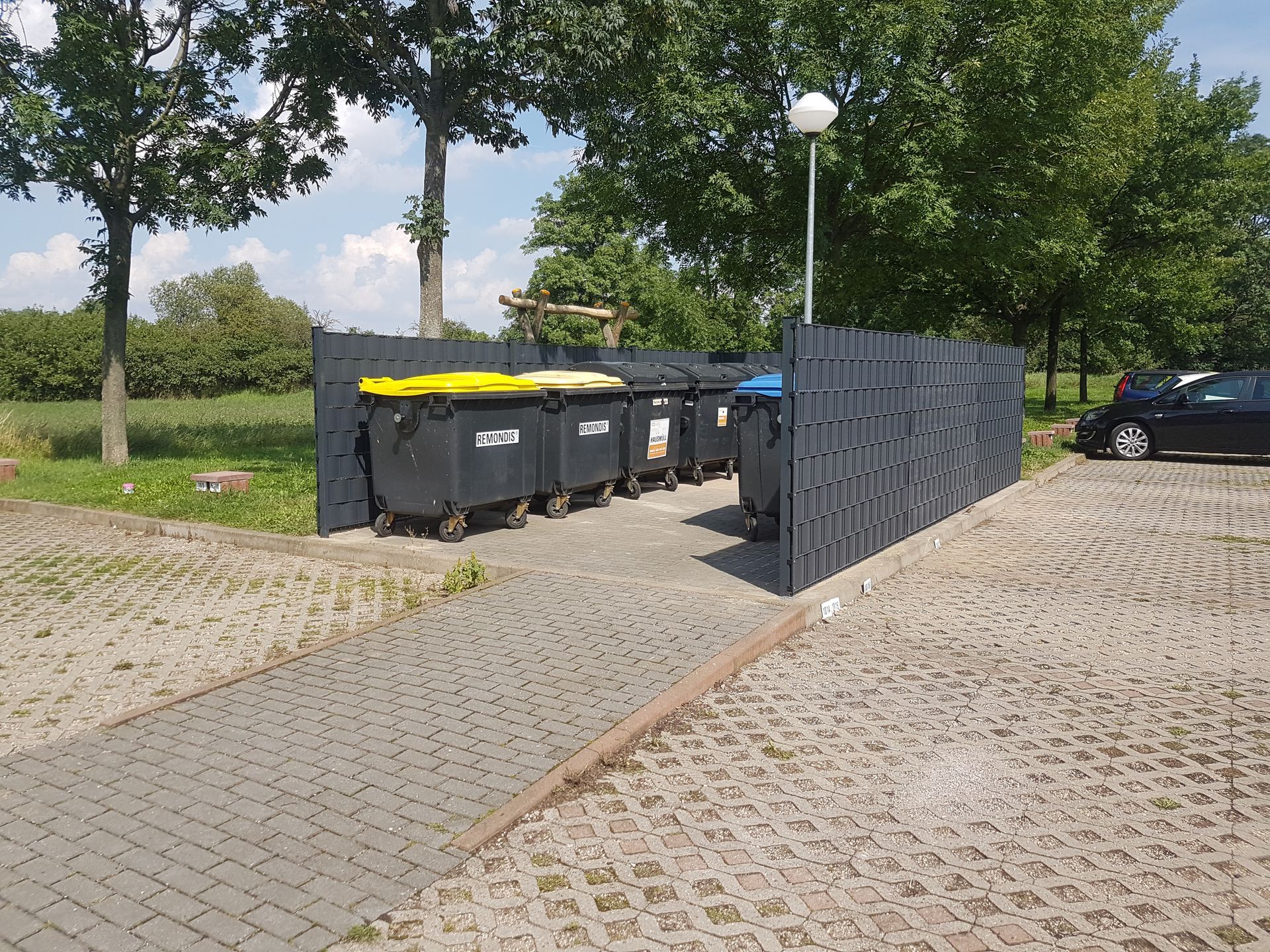 Im Freien stehen Mülleimer neben einem gepflasterten Weg und einem Zaun in einem grasbewachsenen Park, in dessen Nähe Bäume und geparkte Autos stehen.