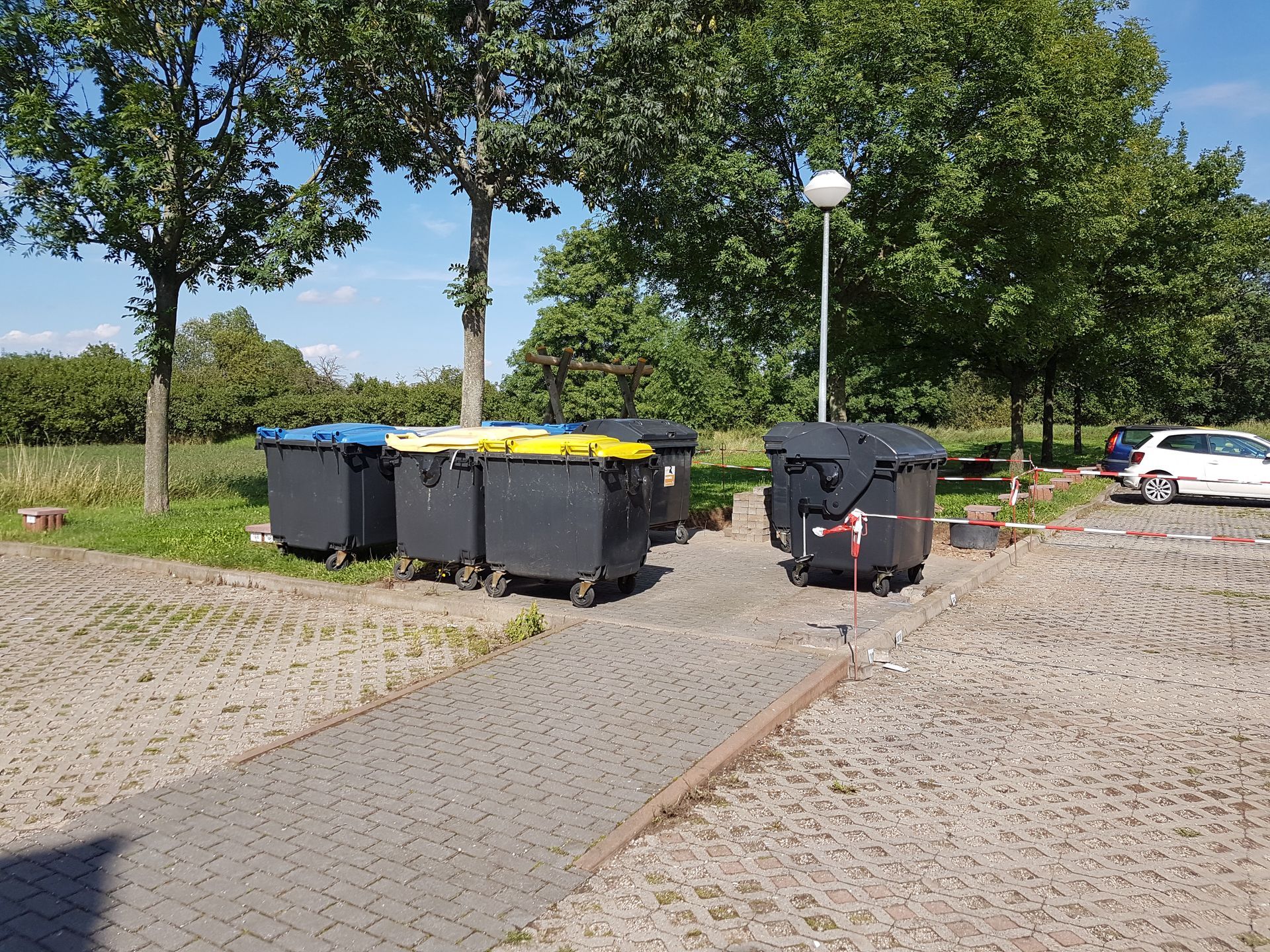 Mülltonnen entlang eines gepflasterten Weges in einem grünen Park, im Hintergrund Bäume und ein geparktes Auto.