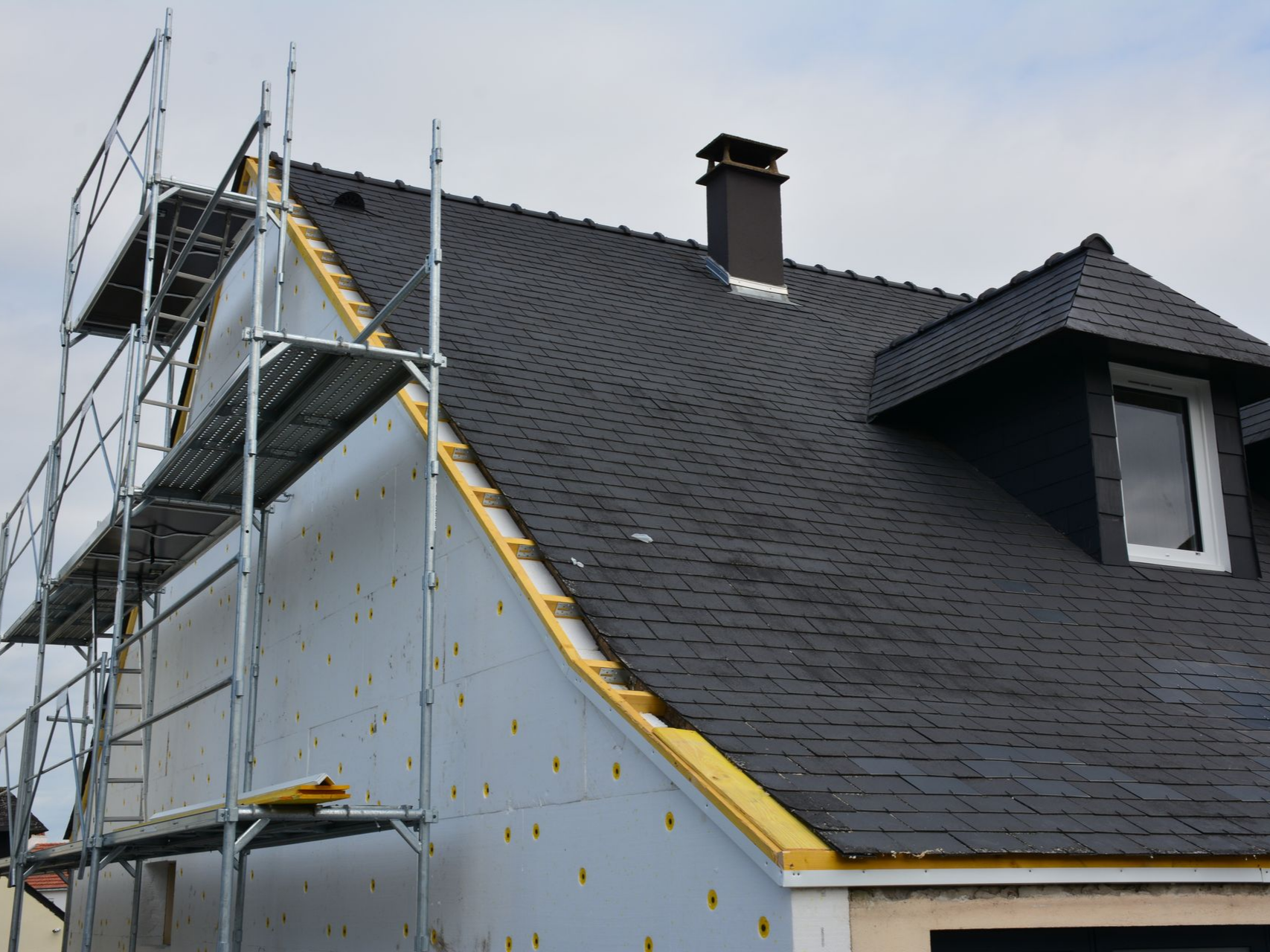 Un échafaudage métallique est adossé au flanc d'une maison au toit pentu de bardeaux sombres, avec une cheminée et une lucarne.