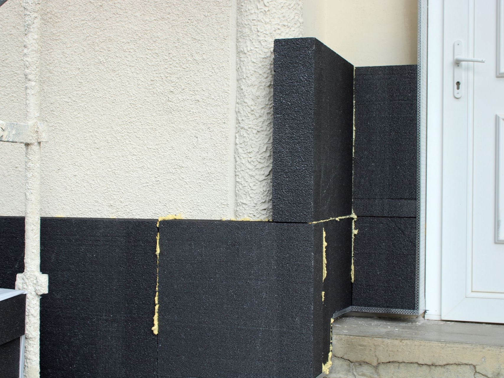 Des panneaux d'isolation noirs sont installés sur le mur extérieur d'un bâtiment, près d'une porte, avec un mastic adhésif jaune.