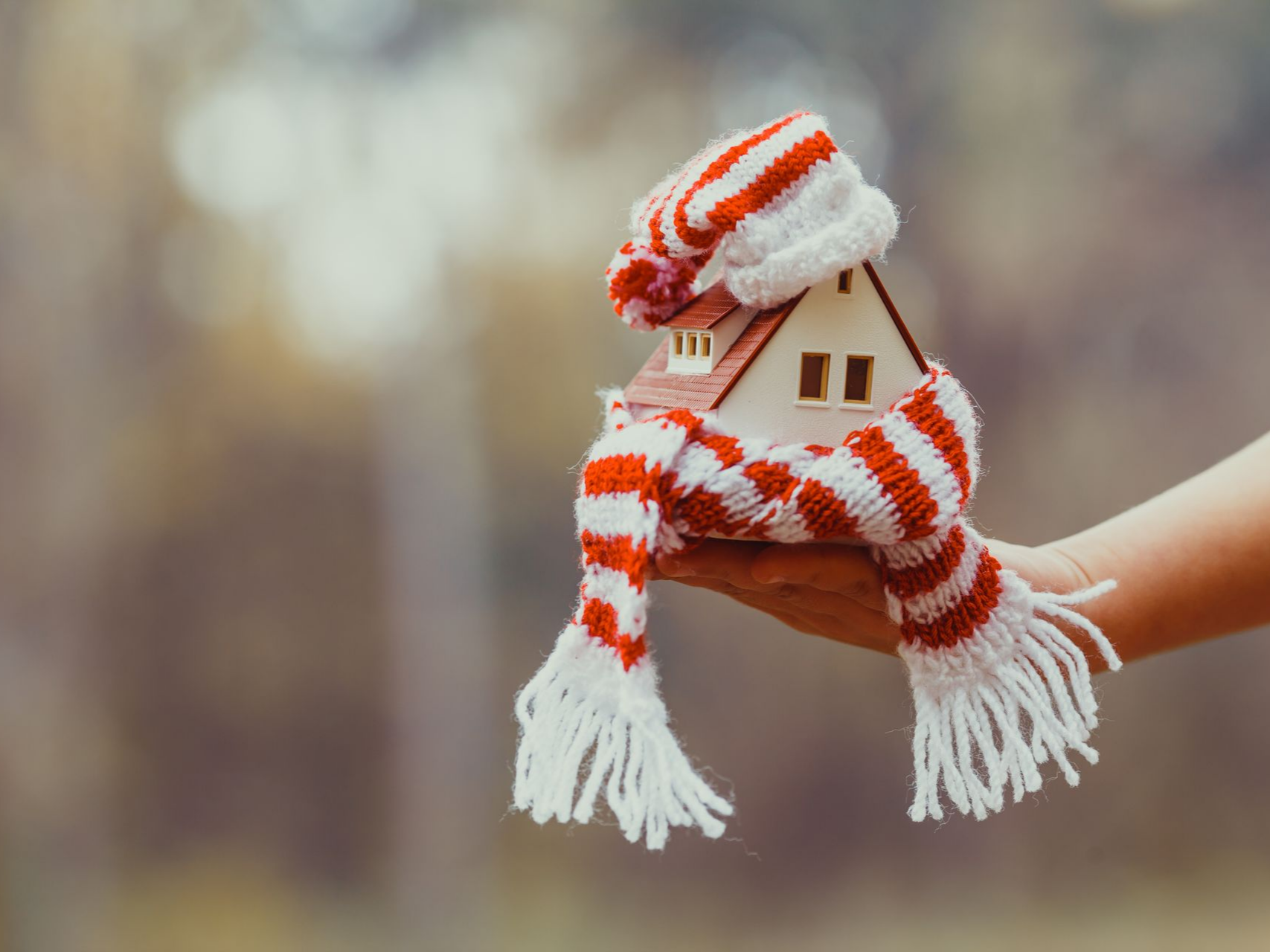 Une main tenant une maison miniature enveloppée dans une écharpe et un bonnet rouges et blancs, évoquant la chaleur.