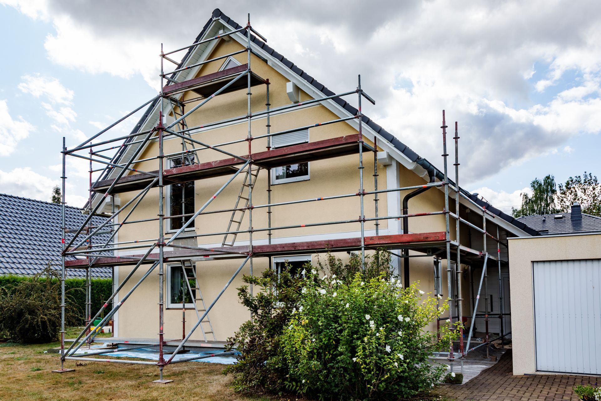 Une maison beige à deux étages, avec un toit à pignon, en cours de travaux d'entretien extérieur, entourée de hauts échafaudages métalliques.