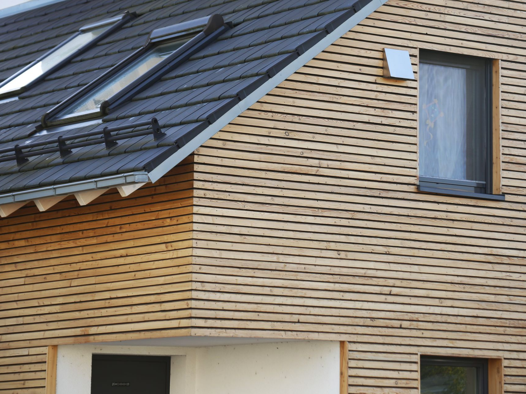 L'extérieur d'une maison moderne présente un bardage horizontal en bois, un toit de tuiles sombres avec des puits de lumière et une porte d'entrée sombre en retrait.
