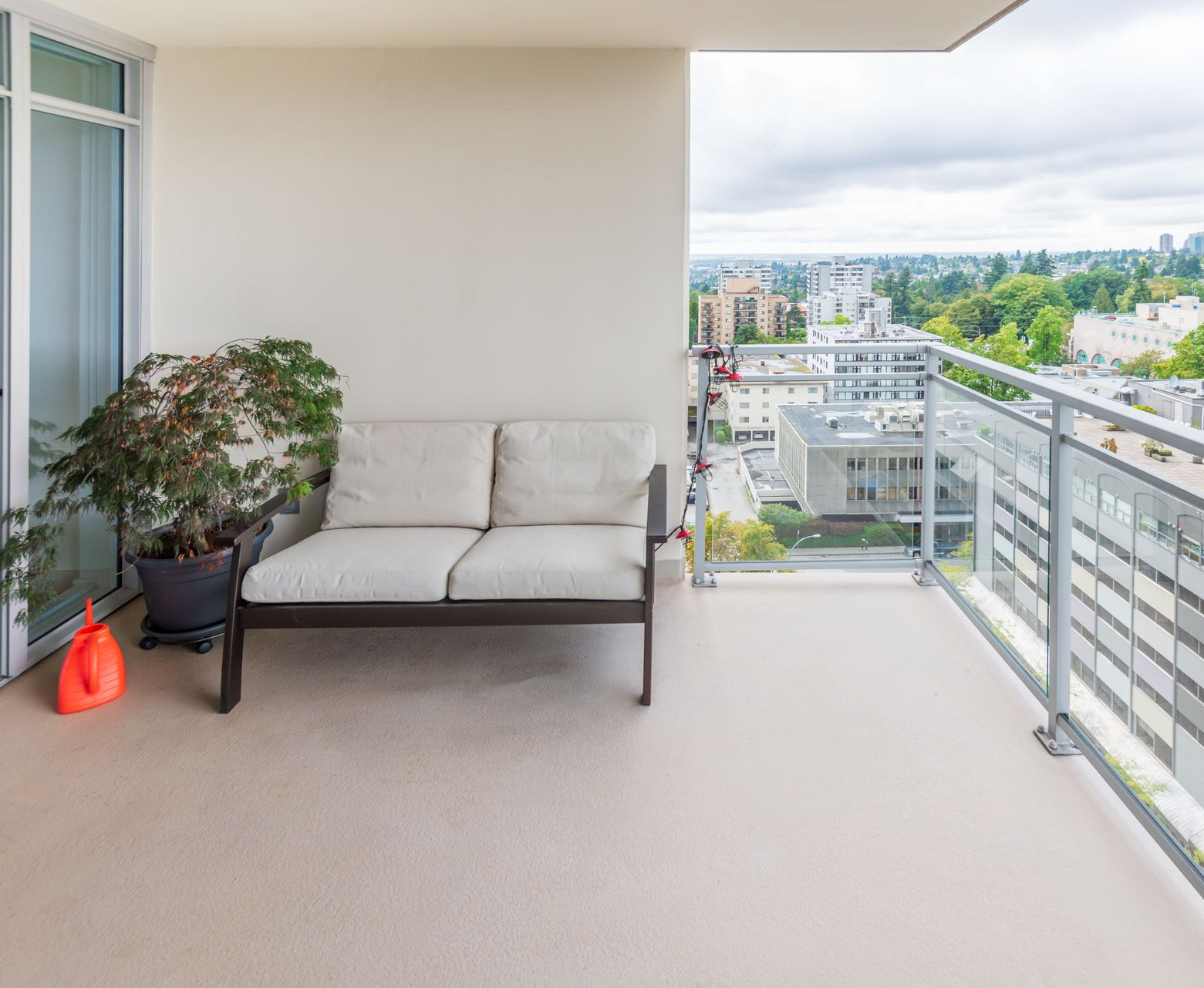 Un balcon avec un canapé deux places blanc cassé, une plante en pot et un arrosoir orange, donnant sur un paysage urbain.