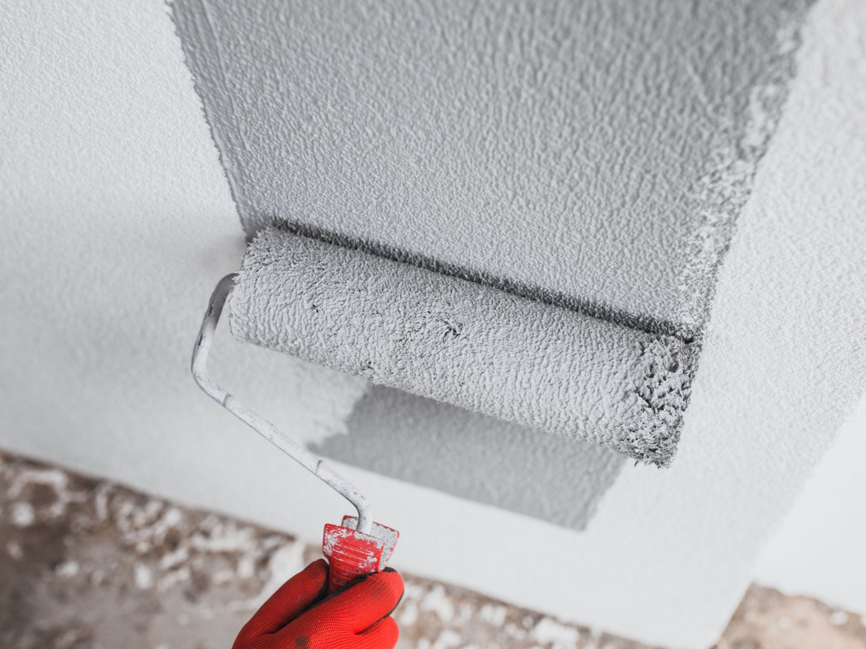 Une main gantée utilise un rouleau à peinture pour appliquer une couche de peinture grise sur un mur.
