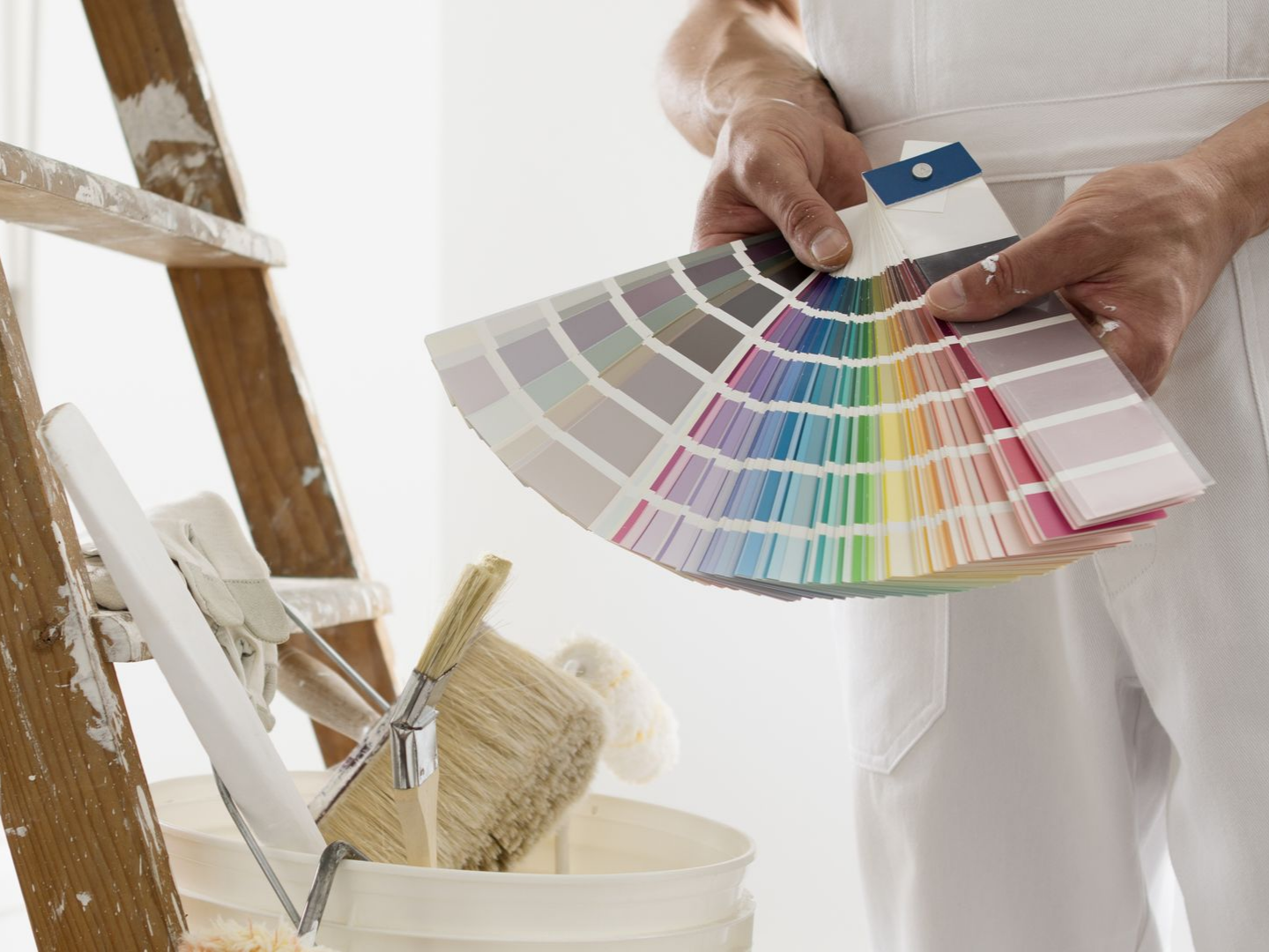 Une personne en salopette blanche tient un éventail d'échantillons de peinture près d'une échelle et d'un seau de peinture.