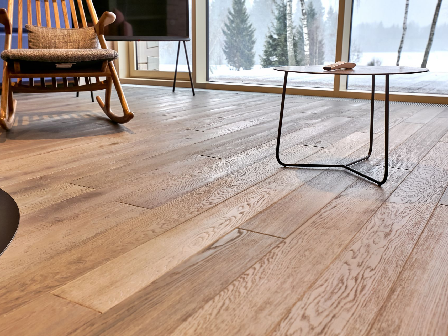 Parquet clair dans une pièce avec un fauteuil à bascule, une petite table et une fenêtre donnant sur un paysage enneigé.