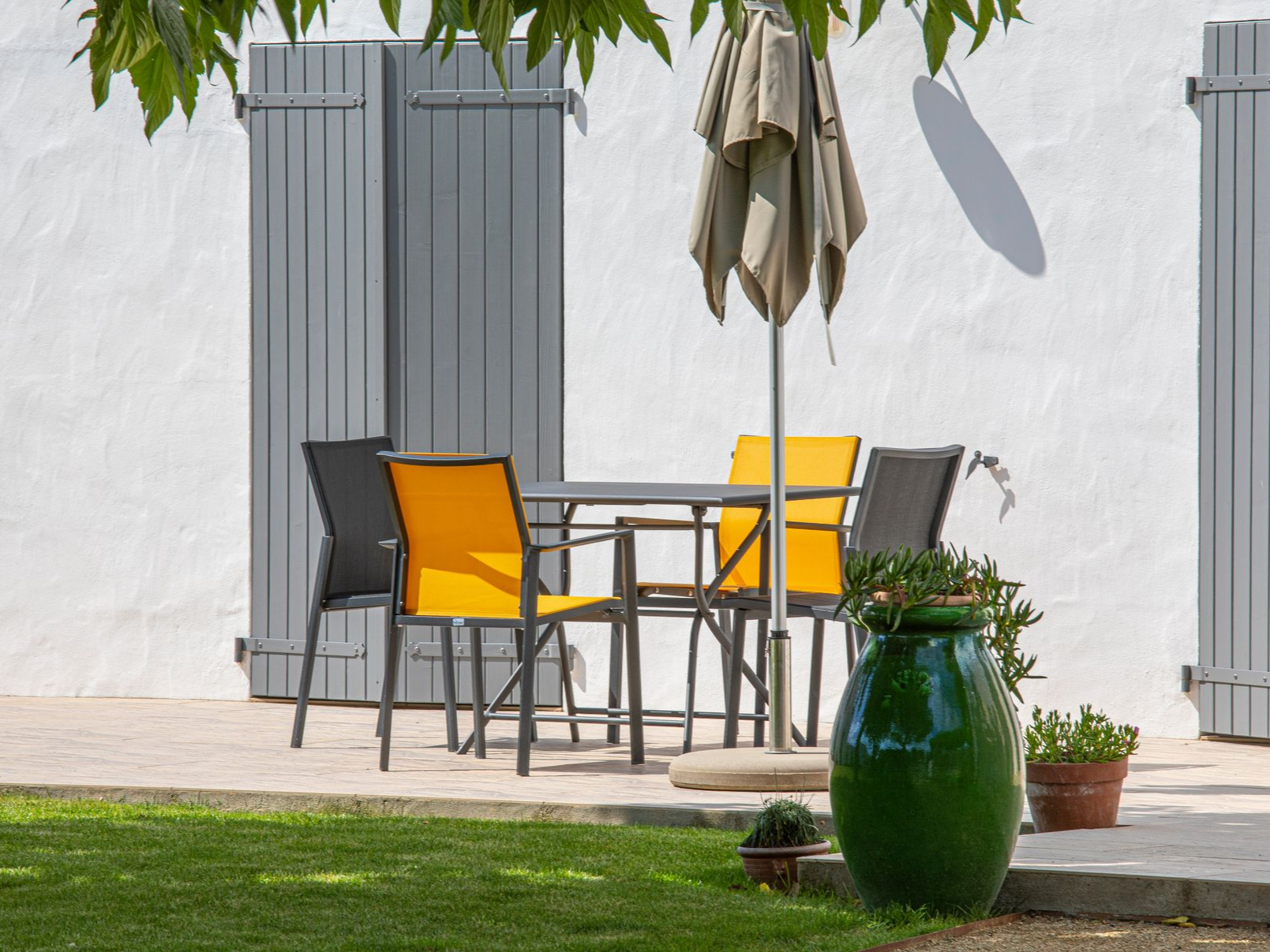 Terrasse avec table et chaises sous un parasol, adossée à un mur en stuc blanc avec des volets gris.