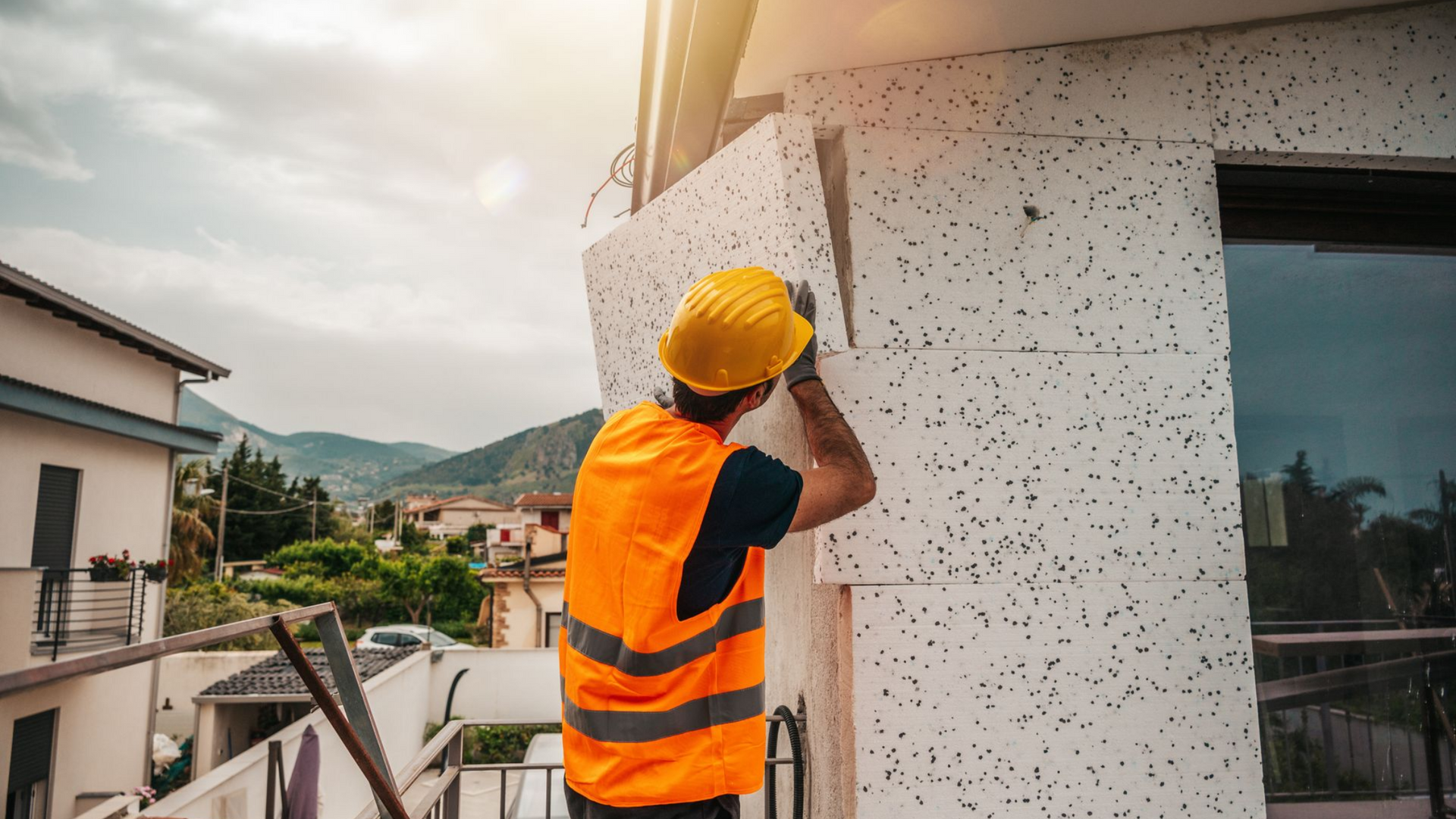 Ouvrier du bâtiment portant un casque jaune et un gilet orange, installant de l'isolant sur la façade d'un bâtiment.