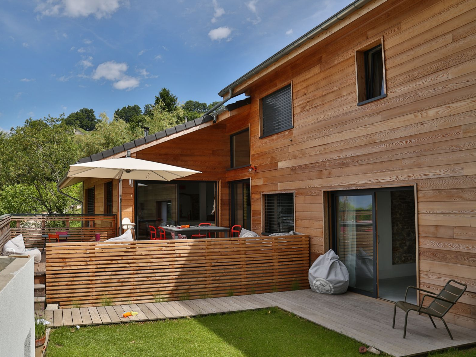 Une maison moderne en bois avec terrasse, parasol et clôture à claire-voie, adossée à une colline verdoyante sous un ciel bleu ensoleillé.