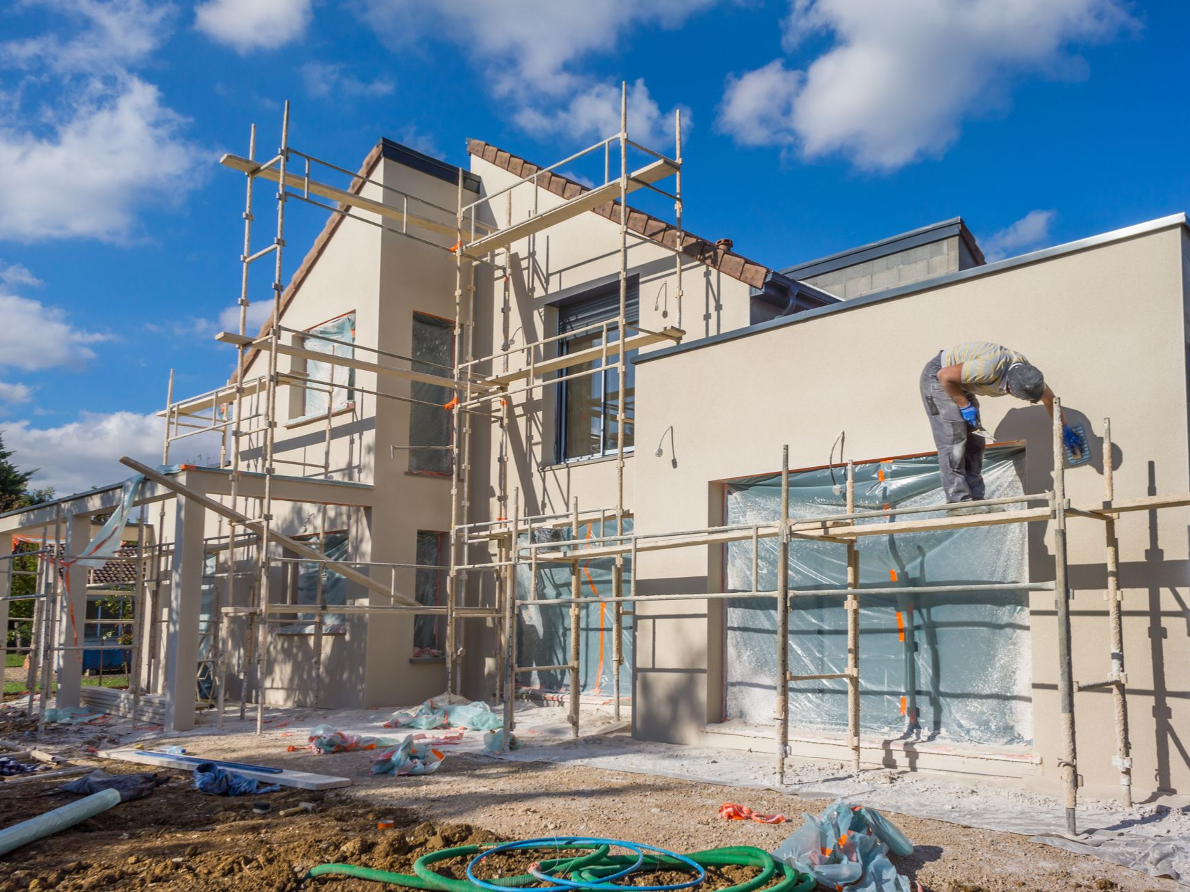 Un ouvrier du bâtiment travaille sur un échafaudage à l'extérieur d'une maison claire sous un ciel d'un bleu éclatant.