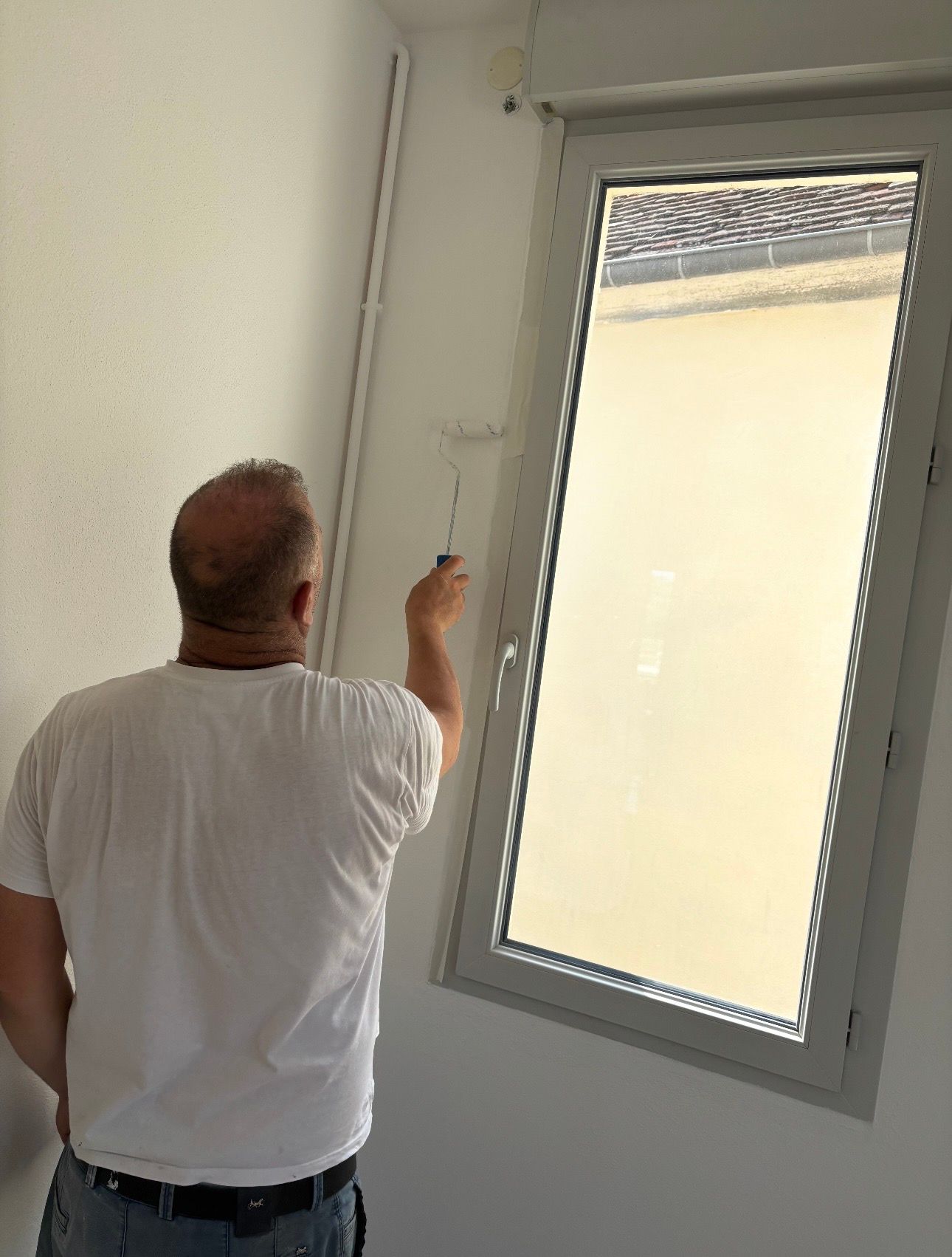 Une personne vêtue d'un t-shirt blanc utilise un rouleau à peinture pour peindre le mur intérieur autour d'une fenêtre.