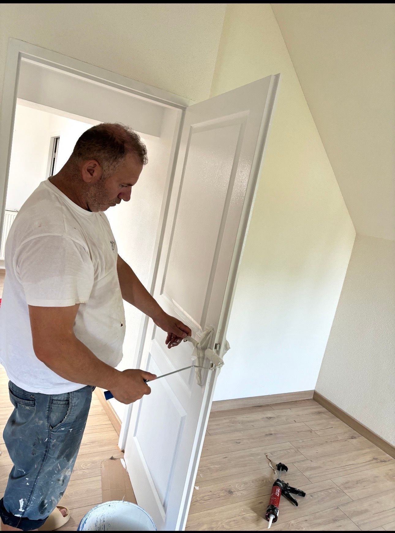 Un peintre en chemise blanche et jean applique de la peinture blanche au rouleau sur une porte en bois dans une pièce au parquet clair.