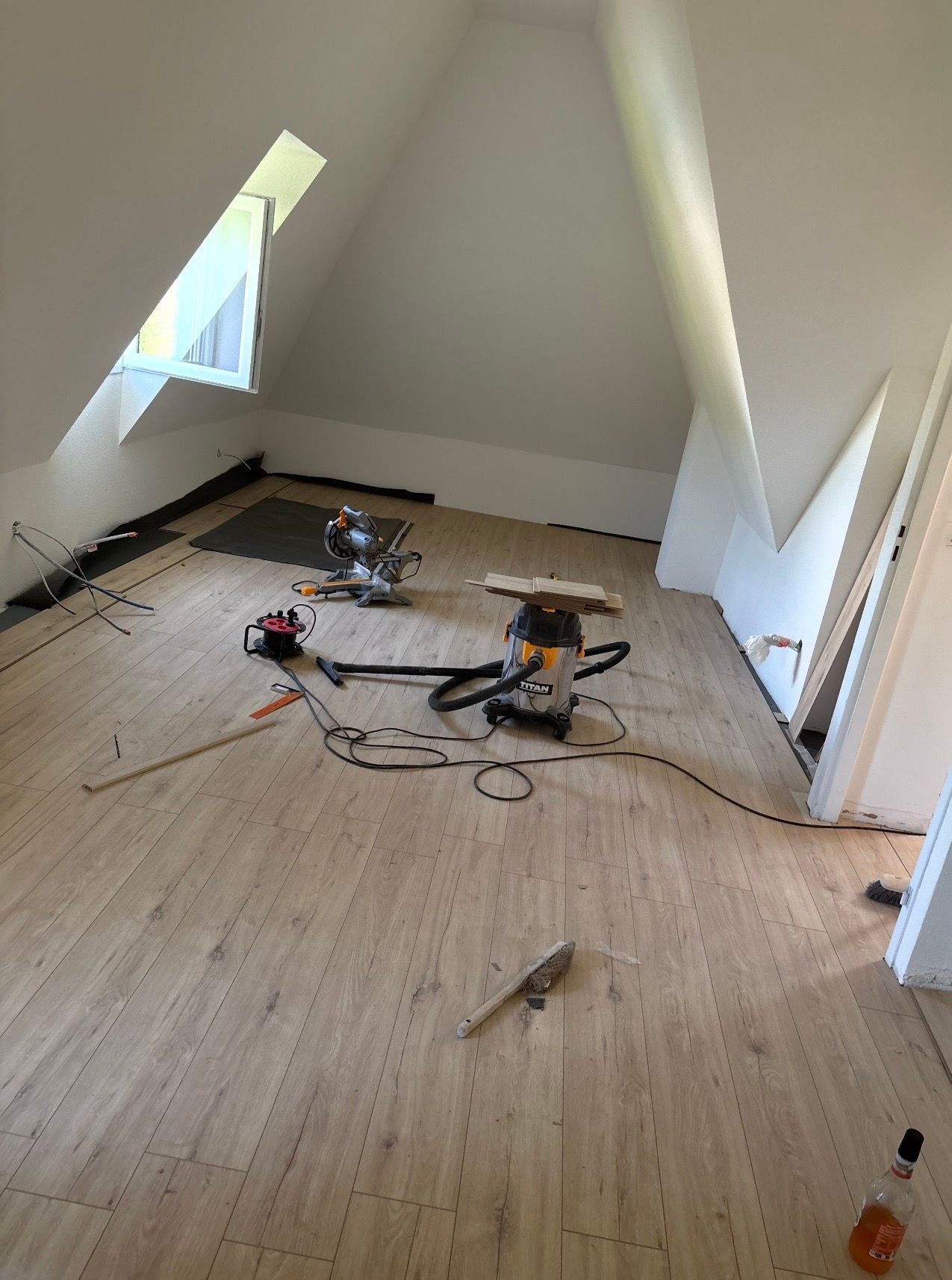 Une pièce mansardée inachevée avec un sol imitation bois, des outils électriques et un aspirateur au centre.