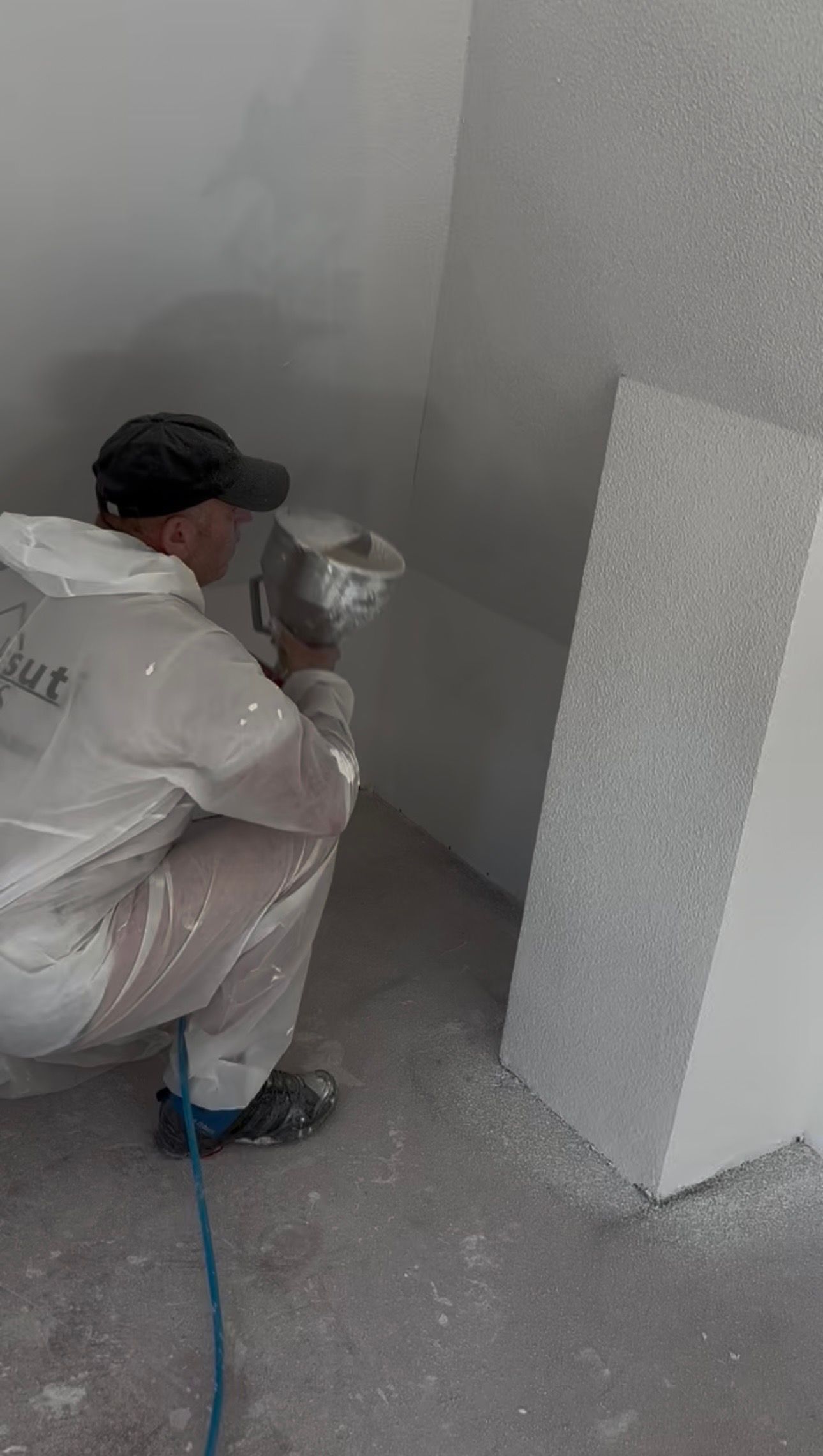 Une personne vêtue d'une combinaison de protection utilise un pistolet à peinture pour appliquer une finition blanche texturée sur un coin de mur.