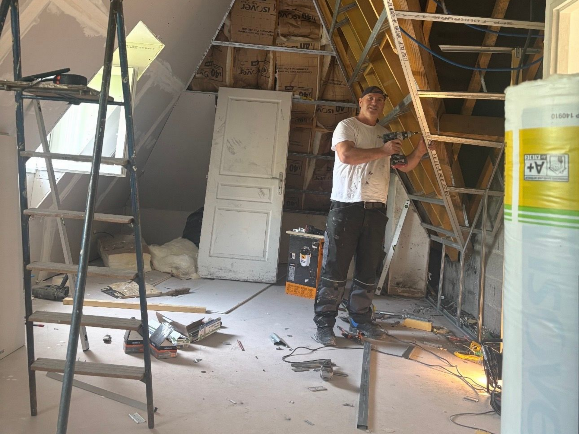 Un ouvrier en t-shirt blanc et pantalon foncé pose avec une perceuse électrique dans un grenier en cours de rénovation.