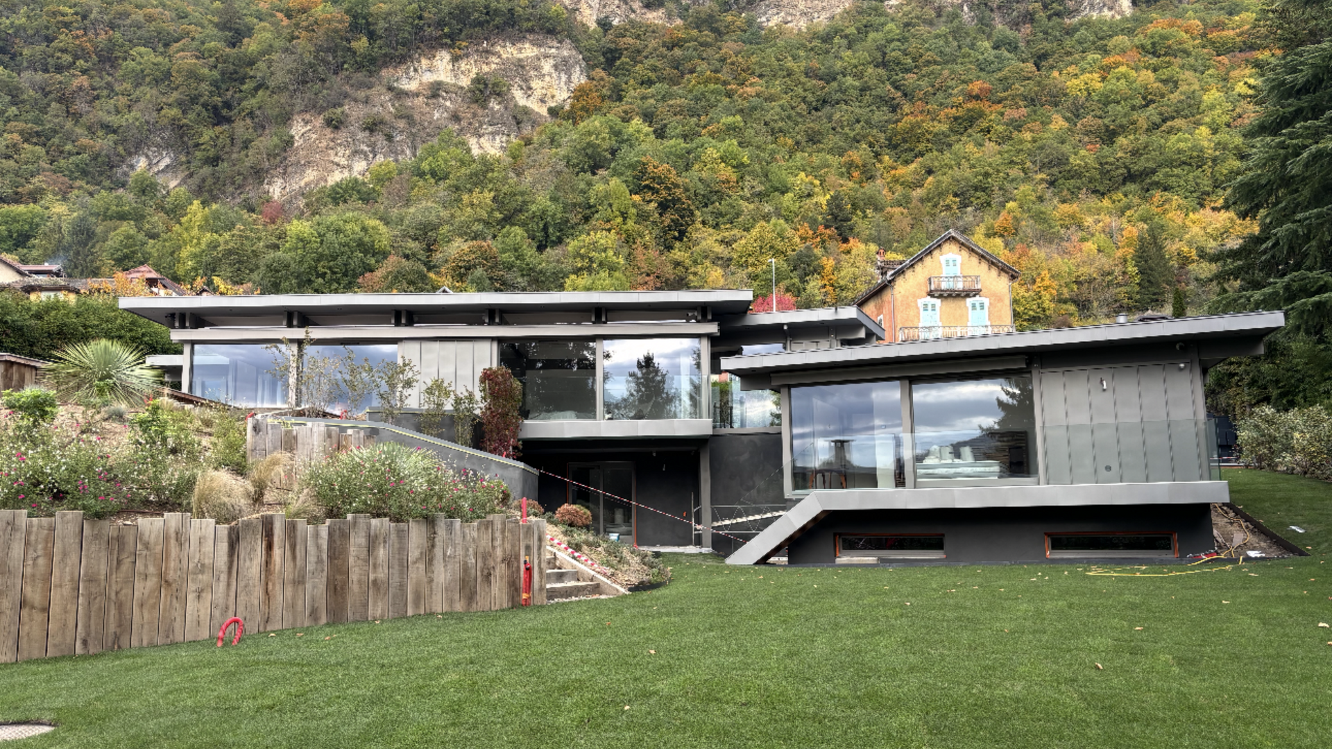 Maison moderne avec de grandes fenêtres, adossée à un paysage montagneux avec pelouse verdoyante et arbres.