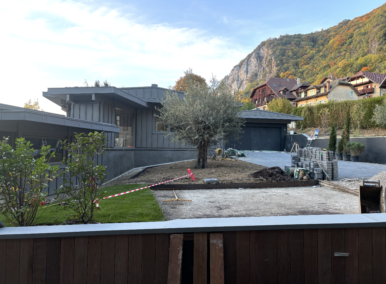 Maison grise moderne avec allée de gravier, olivier et montagnes en arrière-plan.