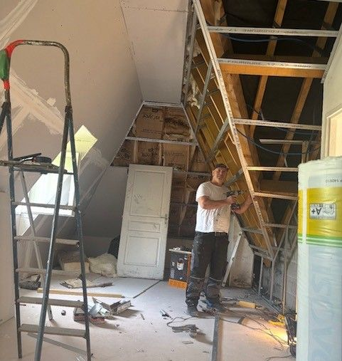 Un homme se trouve dans une pièce en rénovation. La charpente métallique et l'isolation sont visibles. Une échelle, une porte et des outils sont éparpillés autour de lui.