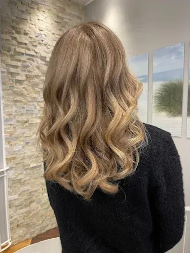 Blonde Frau mit welligem Haar und dunklem Pullover vor einer strukturierten Wand.