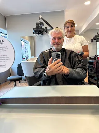 Mann lässt sich im Salon die Haare schneiden und macht ein Spiegel-Selfie. Friseur lächelt hinter ihm.