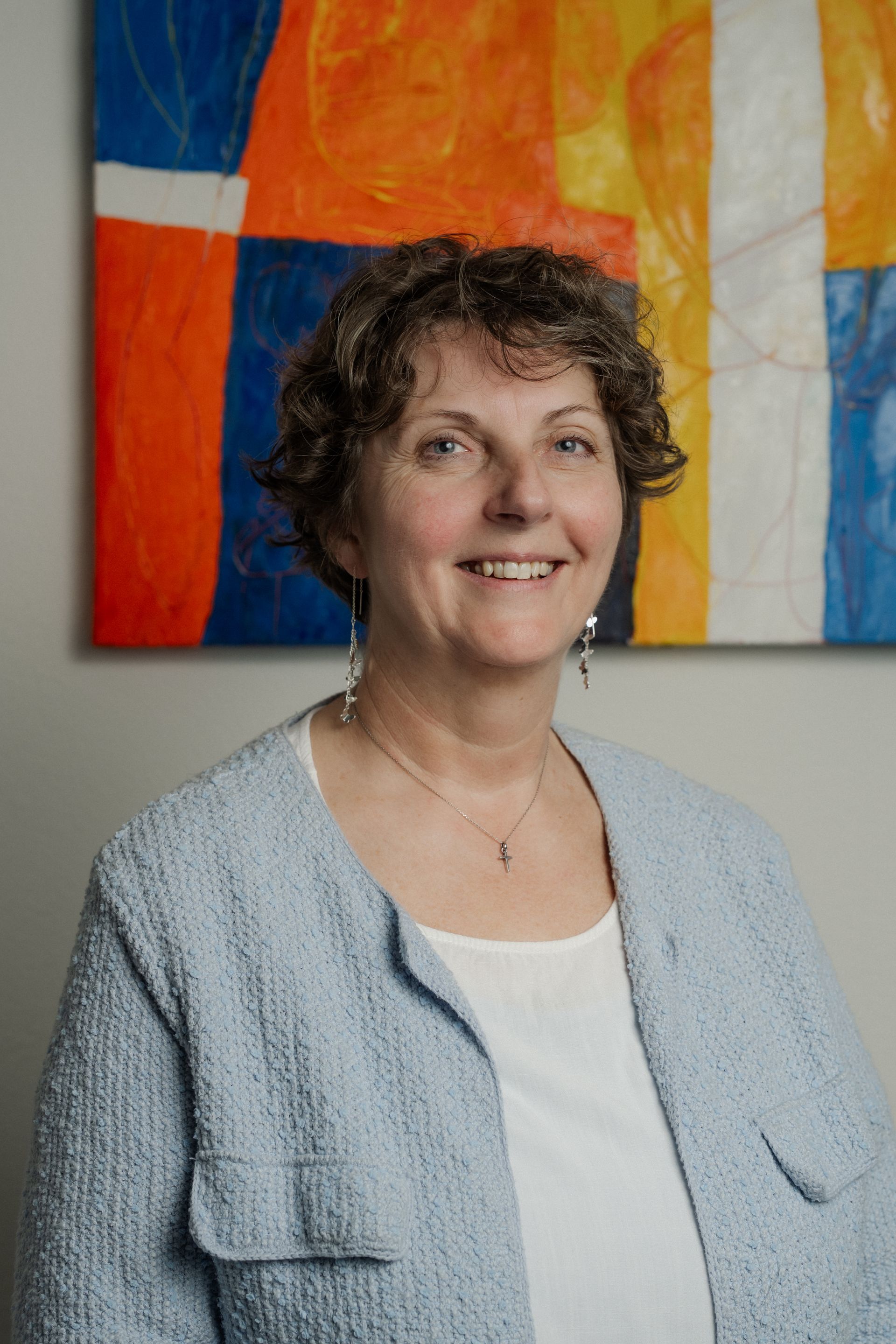 Eine Frau in einer hellblauen Jacke lächelt vor farbenfroher abstrakter Kunst.