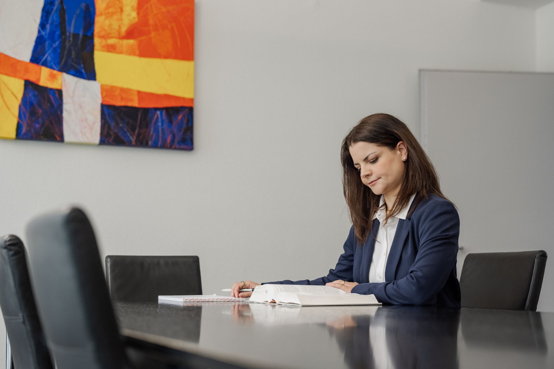 Frau im Blazer sitzt an einem Tisch und liest ein Buch. Modernes Büroambiente, Kunstwerke an der Wand.