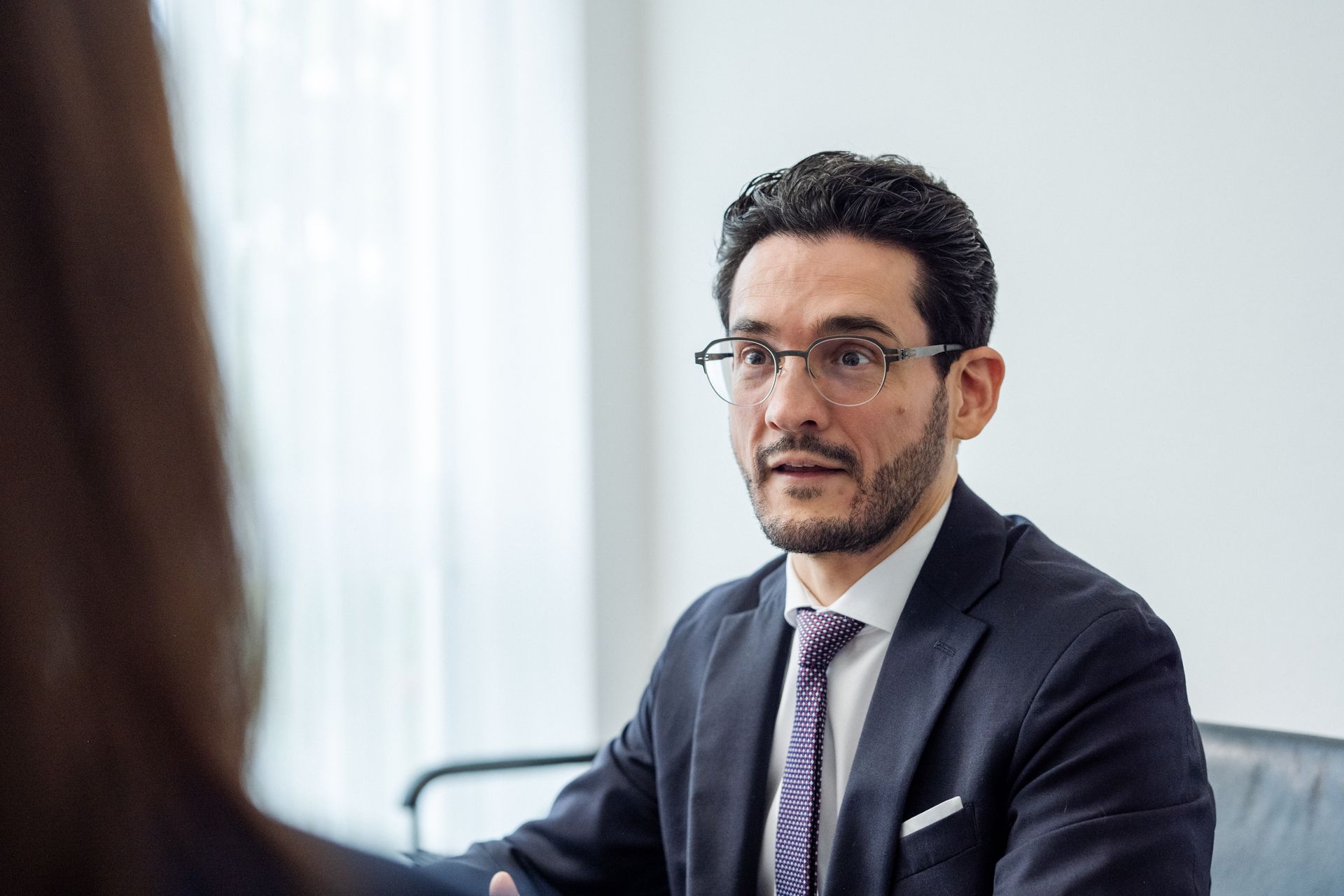 Ein Mann im Anzug und mit Brille unterhält sich in einem hellen Büro mit jemandem außerhalb des Bildausschnitts.