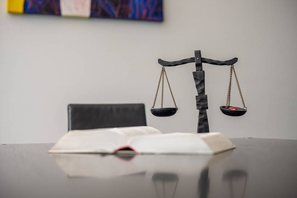 Ein aufgeschlagenes Buch und eine Waage liegen auf einem dunklen Tisch, davor ein Stuhl und ein Wandbild.