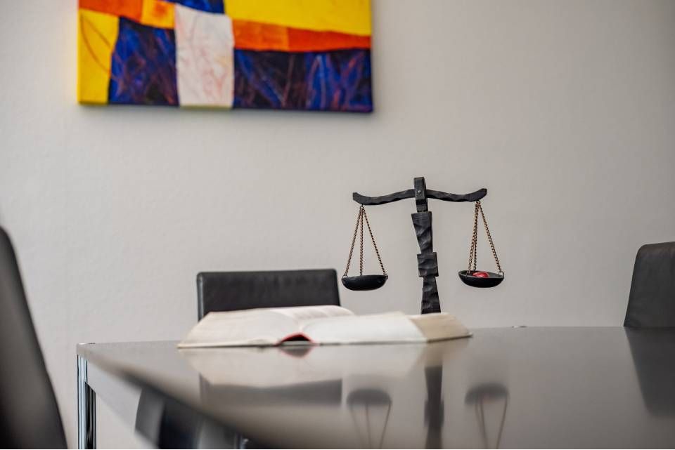 Eine Justitiawaage und ein aufgeschlagenes Buch auf einem dunklen Tisch, an der Wand ein farbenfrohes Gemälde.