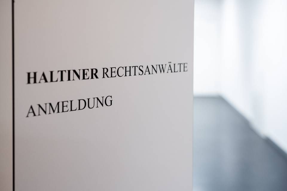 Schild mit der Aufschrift "HALTINER RECHTSANWÄLTE ANMELDUNG" in schwarzer Schrift auf weisser Wand.