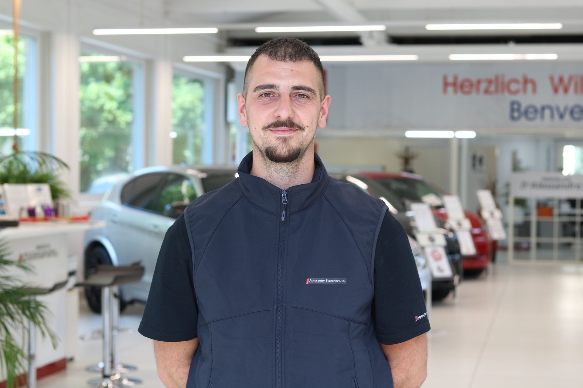 Person mit kurzen schwarzen Haaren, die ein schwarzes Hemd trägt und vor einem einfarbig weissen Hintergrund lächelt