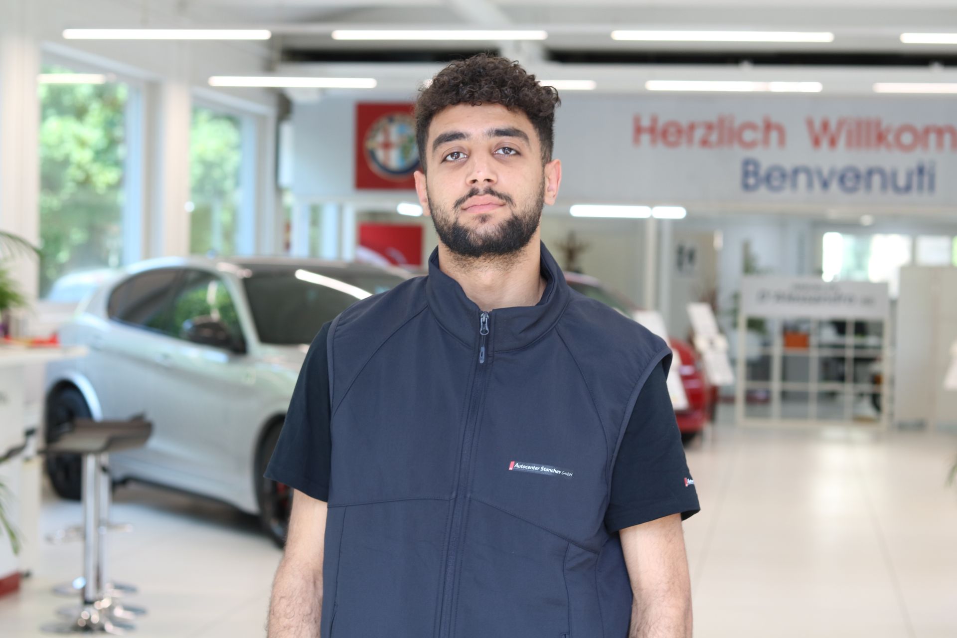 Person mit kurzen schwarzen Haaren, die ein schwarzes Hemd trägt und vor einem einfarbig weissen Hintergrund lächelt