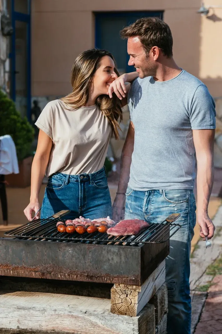 Una pareja está asando carne en una barbacoa al aire libre. La mujer sonríe, mirando al hombre.