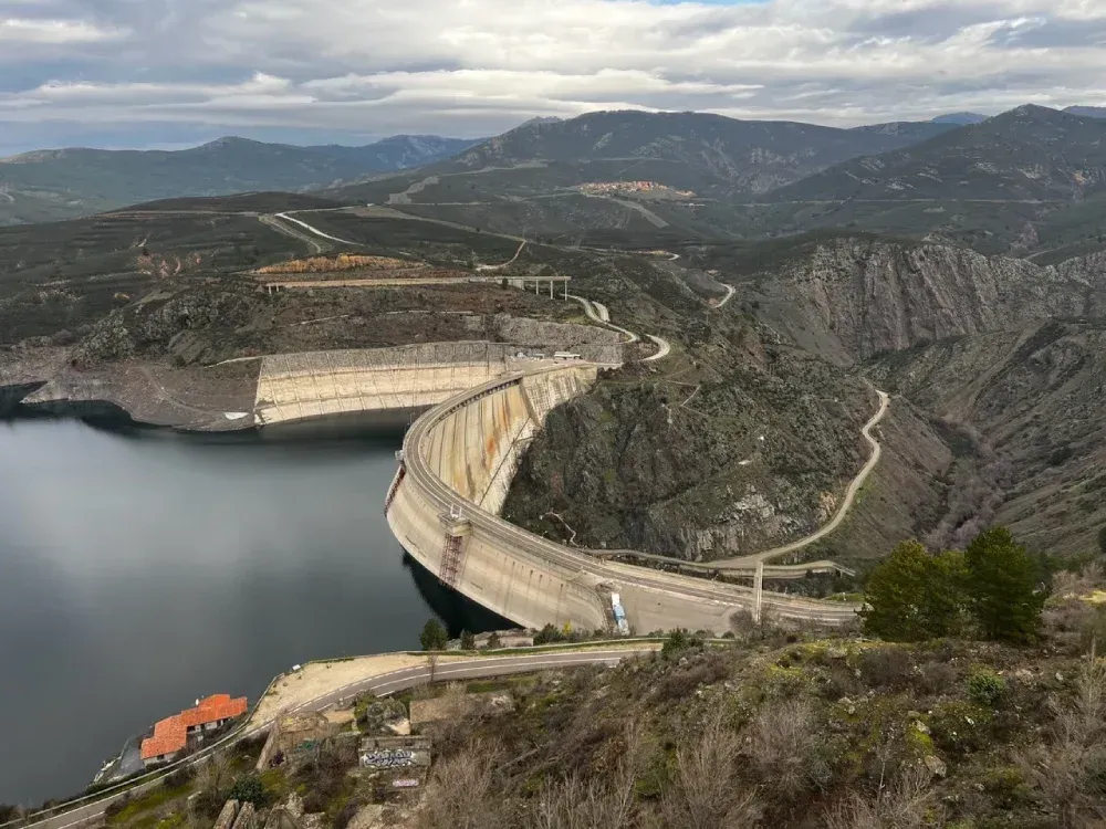 Presa curva de hormigón que cruza un desfiladero, creando un gran embalse. Montañas y carreteras sinuosas al fondo.