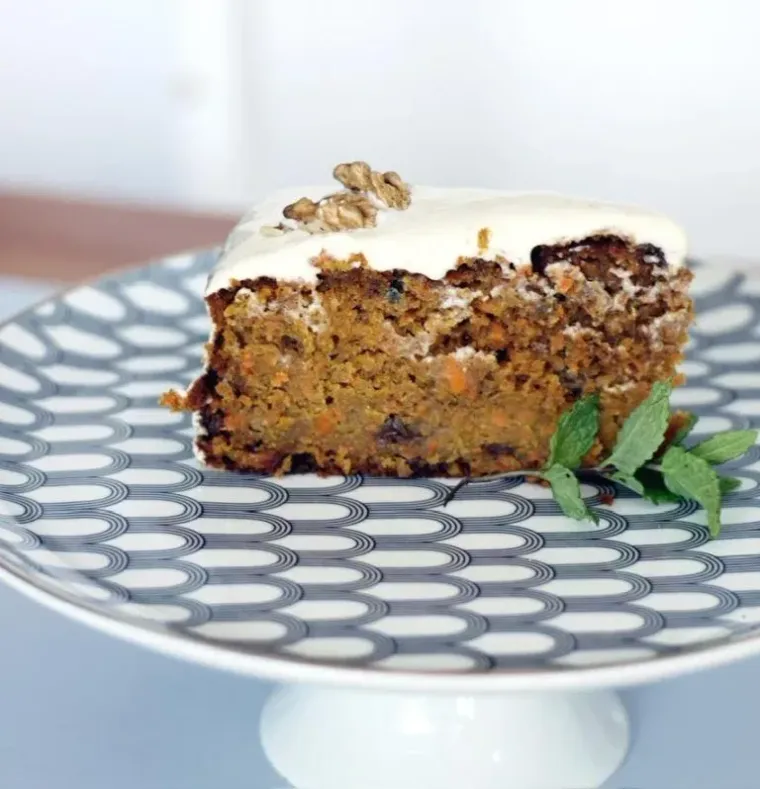 Rebanada de pastel de zanahoria con glaseado blanco y una nuez, servida en un plato decorado con menta.