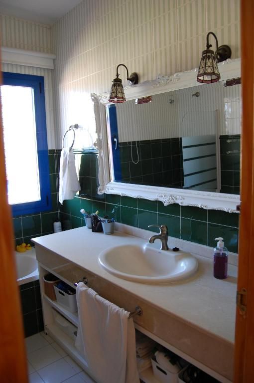 Baño con lavabo blanco, espejo y pared de azulejos verdes; ventana con marco azul.