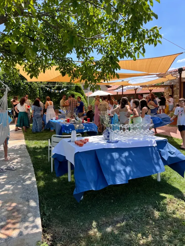 Fiesta al aire libre con gente reunida alrededor de mesas. Manteles azules y blancos, sombrillas y césped verde.