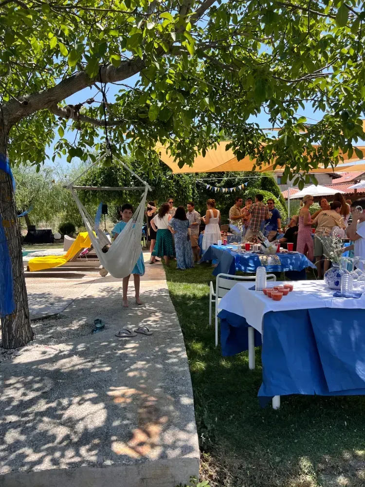 Persona en una hamaca bajo un árbol, fiesta con mesas cubiertas con manteles azules, al aire libre.