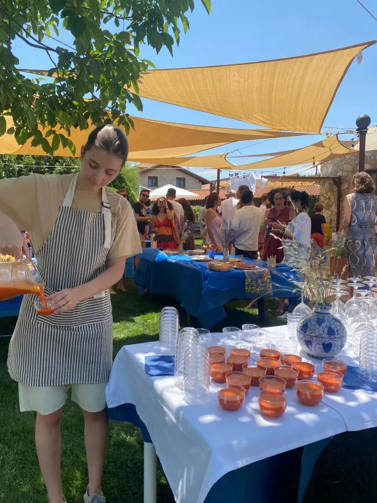 Una persona con delantal sirve una bebida en una reunión al aire libre; otras personas se ven al fondo.
