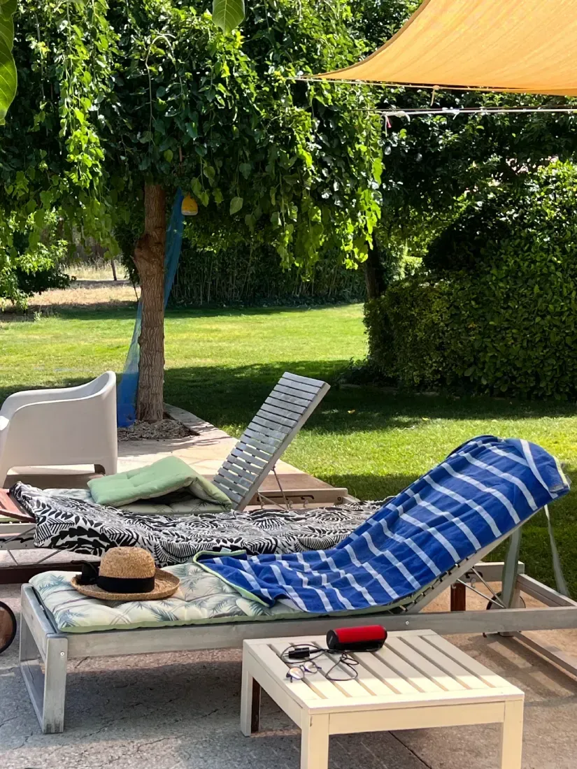 Tumbonas en un patio bajo una sombrilla, con sombrero, toalla y mesita; césped verde y árboles al fondo.
