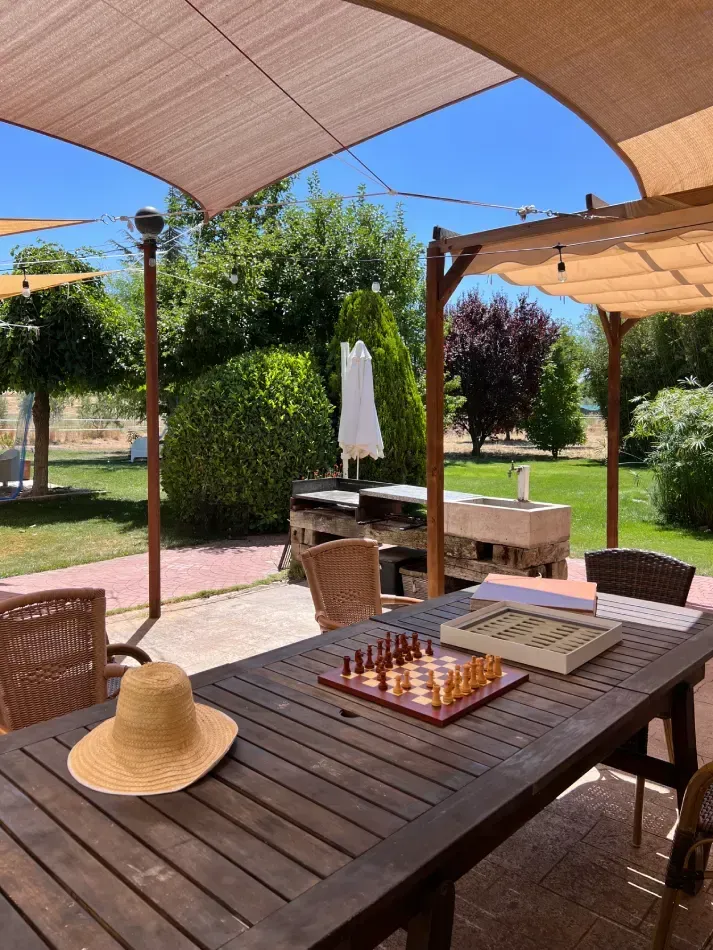 Mesa exterior con juegos de mesa y sombrero de paja, a la sombra de un toldo color canela, con vistas a un césped verde.