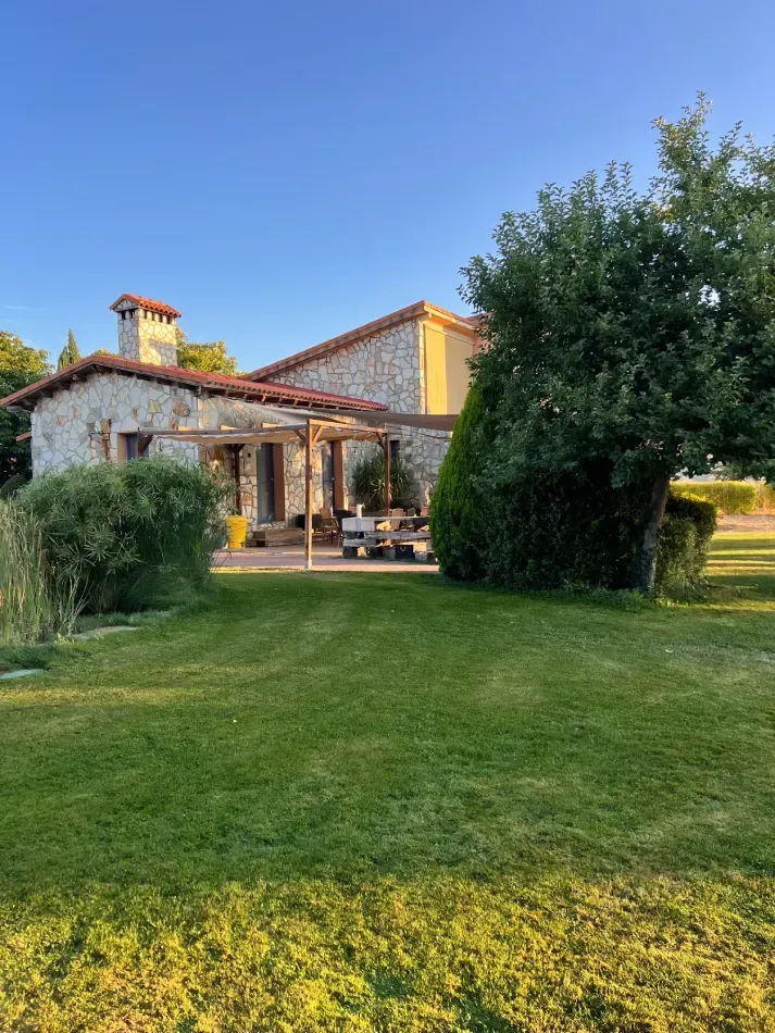 Casa de piedra con patio, césped verde y árboles bajo un cielo azul despejado.