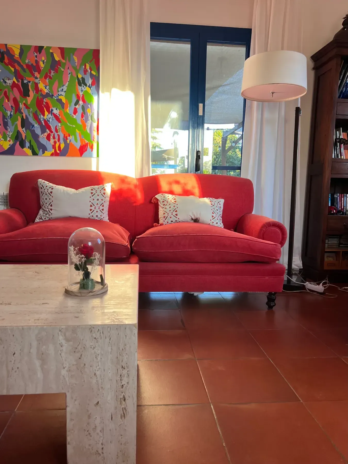 Sofá rojo con cojines blancos, obras de arte y una mesa de centro en una habitación iluminada por el sol.