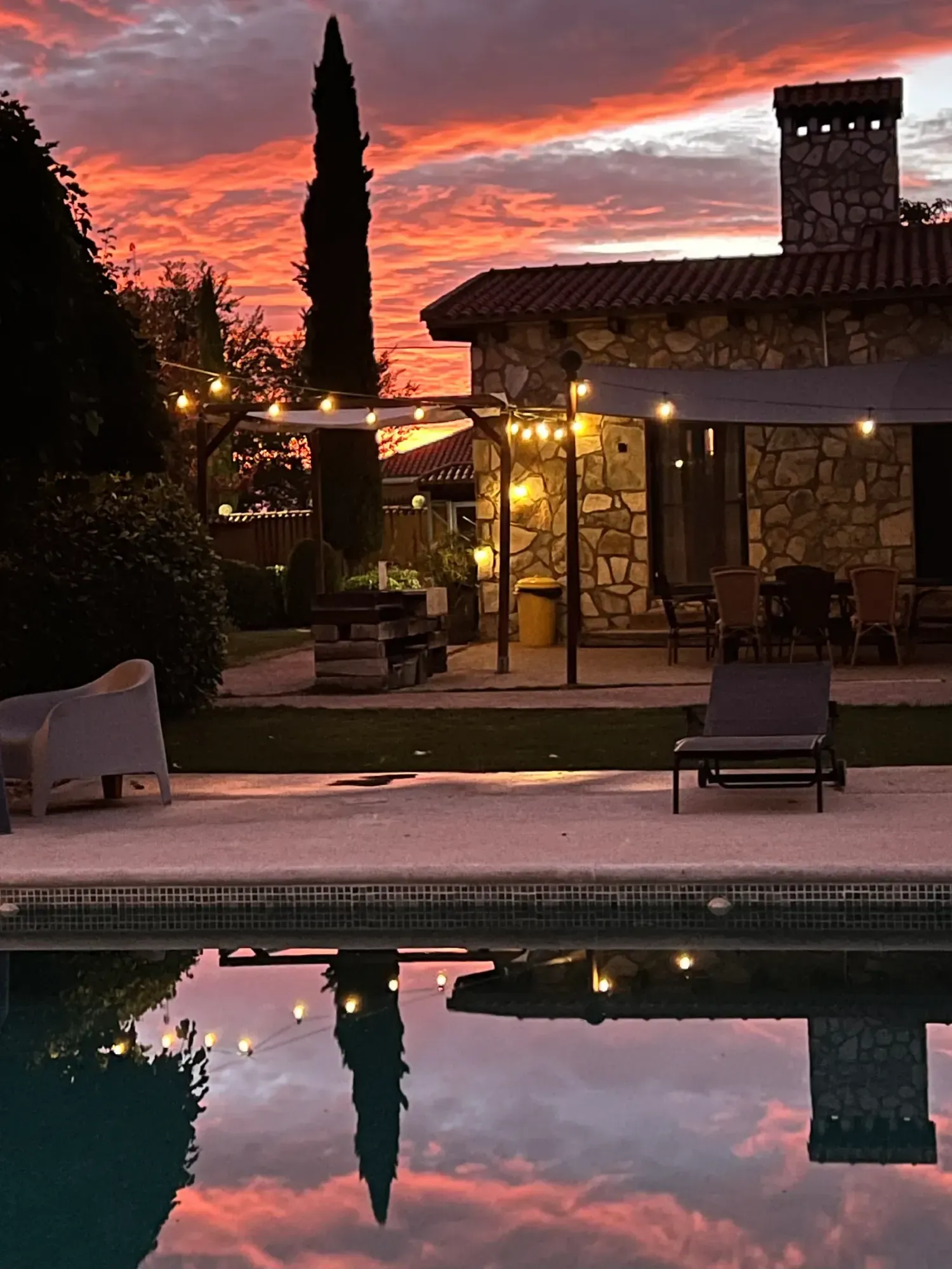 Atardecer sobre una casa de piedra con piscina. Las luces se reflejan en el agua. Cielo rosa y naranja.