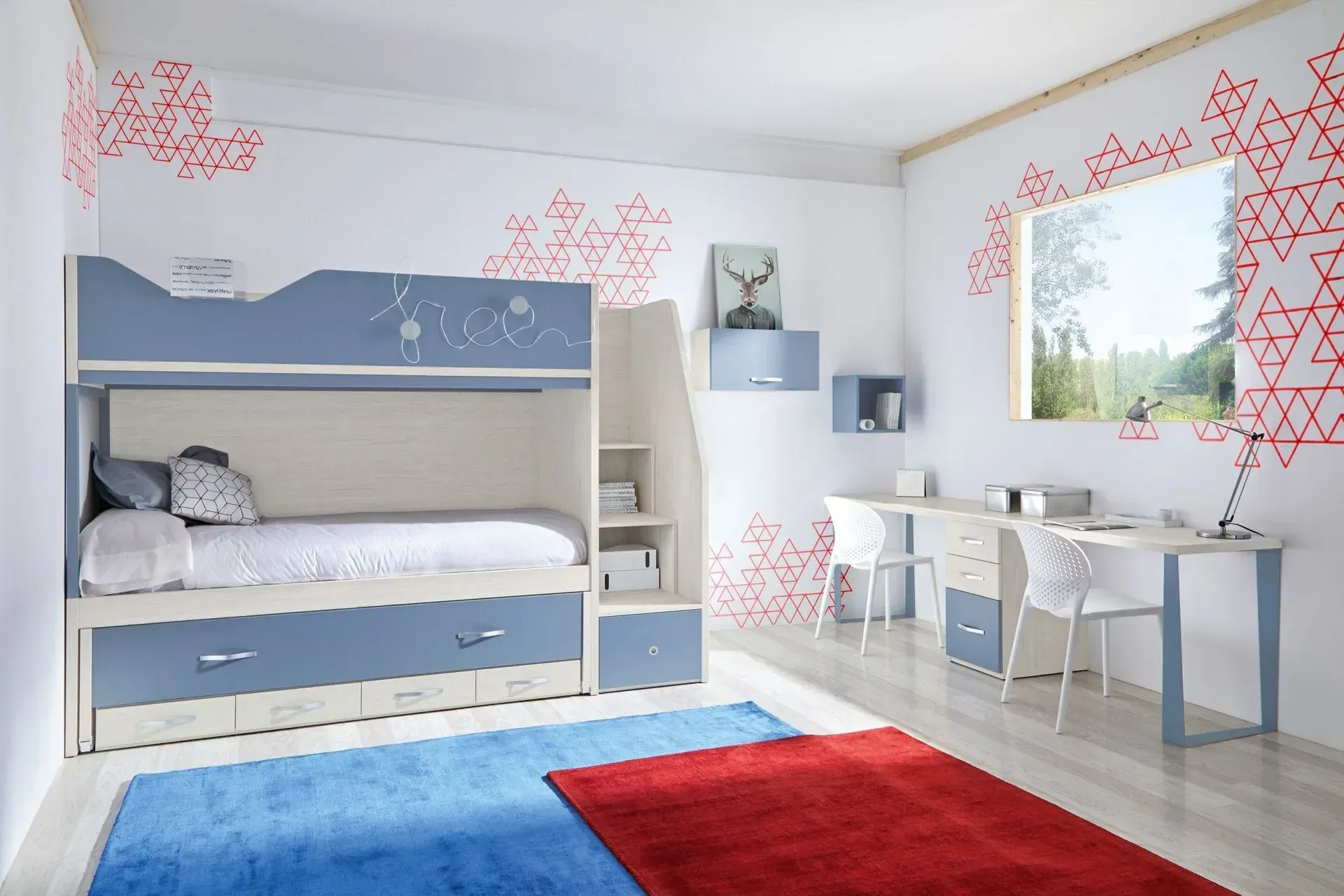 Litera azul y blanca en el dormitorio de un niño con dos escritorios, una alfombra roja y azul y una decoración de pared geométrica roja.